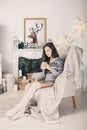 Beautiful girl near fireplace with Christmas tree, candles sits in comfortable armchair. Scandinavian cozy home decor Royalty Free Stock Photo