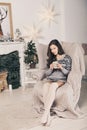 Beautiful girl near fireplace with Christmas tree, candles sits in comfortable armchair. Scandinavian cozy home decor Royalty Free Stock Photo