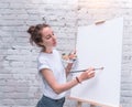 Beautiful girl paints picture, artist holds color palette in hand, brush. White blank canvas, easel. Summer in city Royalty Free Stock Photo