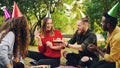 Beautiful girl in party hat is celebrating birthday with friends in park on picnic making wish, blowing candles on cake