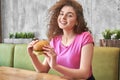 Girl sitting in cafe, holding tasty hamburger, smiling. Royalty Free Stock Photo