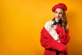 A beautiful girl in a red coat and beret stands on a yellow background with horror, looks away pointing finger there