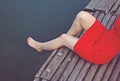 Beautiful girl in red dress is sitting on the wooden pier. Woman is dangling her feet into the lake. Rustic and natural photo