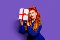 A beautiful girl with red hair and red lipstick in a blue dress in the style of pin-up on a purple background holds a gift and Royalty Free Stock Photo