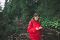 Beautiful girl in red raincoat uses smartphone on a background of mountain stream and forest. Hiker woman watching map and Royalty Free Stock Photo