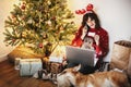 beautiful girl in reindeer antlers holding credit card and shopping on phone, sitting with laptop at golden beautiful christmas t Royalty Free Stock Photo