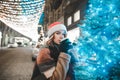 Beautiful girl in santa hat stands near Christmas tree with Christmas lights on the background of the decorated street, looks in Royalty Free Stock Photo