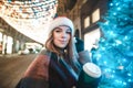 Beautiful girl in a Santa hat stands near a Christmas tree on a street background decorated with Christmas lights, holds a cup of Royalty Free Stock Photo