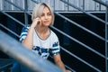 Beautiful girl with short white hair dressed in jeans in urban style sits on the metal stairs. Place for text Royalty Free Stock Photo