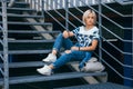 Beautiful girl with short white hair dressed in jeans in urban style sits on the metal stairs. Place for text Royalty Free Stock Photo