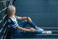 Beautiful girl with short white hair dressed in jeans in urban style sits on the metal stairs. Place for text Royalty Free Stock Photo