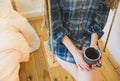 Beautiful girl is sitting on swing in loft minimalist interior. Young woman in men`s checkered shirt is drinking coffee. Cozy Royalty Free Stock Photo