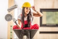 Beautiful girl sitting in a wheelbarrow inside the construction site with building tools in her hands wearing overall and a hard Royalty Free Stock Photo