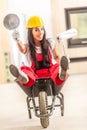 Beautiful girl sitting in a wheelbarrow inside the construction site with building tools in her hands wearing overall and a safety Royalty Free Stock Photo