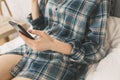 Beautiful girl is sitting on the white bed linen. Young woman in men`s checkered shirt is searching in social network on white Royalty Free Stock Photo