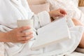 Young woman in white terry robe is drinking coffee and reading magazine or book in bedroom. Close-up Royalty Free Stock Photo