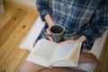 Beautiful girl is sitting on the wooden floor. Young woman in men`s checkered shirt is drinking coffee and reading a book on whit Royalty Free Stock Photo