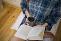 Beautiful girl is sitting on the wooden floor. Young woman in men`s checkered shirt is drinking coffee and reading a book on whit Royalty Free Stock Photo