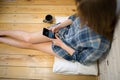 Beautiful girl is sitting on the wooden floor. Young woman in men`s checkered shirt is drinking coffee and searching in social Royalty Free Stock Photo