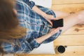 Beautiful girl is sitting on the wooden floor. Young woman in men`s checkered shirt is drinking coffee and searching in social Royalty Free Stock Photo