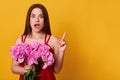 Beautiful girl standing with bouquet of rosy peonies in her hands, pointing blank space with her index finger isolated over yellow Royalty Free Stock Photo