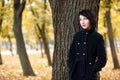 Beautiful girl standing near tree in autumn city park, yellow leaves on background, fall season, dressed in black coat Royalty Free Stock Photo