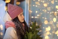 A beautiful girl stands near a decorated window holding a Christmas tree in her hands, dressed in a hat and a warm scarf Royalty Free Stock Photo