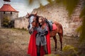 A beautiful girl stands next to a horse against the background of a medieval fortress. Fantasy costume, chain mail, cloak. A woman Royalty Free Stock Photo