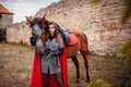 A beautiful girl stands next to a horse against the background of a medieval fortress. Fantasy costume, chain mail, cloak. A woman Royalty Free Stock Photo