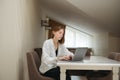 Beautiful girl student in casual clothes sits in the apartment at the desk and works at a laptop. Attractive young female Royalty Free Stock Photo