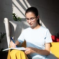 Beautiful girl student writes with a pen an assignment in a notebook while sitting on an armchair in a cafe Royalty Free Stock Photo