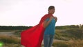 Beautiful girl superhero standing on the field in a red cloak, cloak fluttering in the wind. Slow motion. A young girl