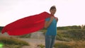 Beautiful girl superhero standing on the field in a red cloak, cloak fluttering in the wind. Slow motion. A young girl