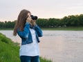 Beautiful girl tourist in a cotton jacket taking photos with a professional camera on the banks of the river in windy weather