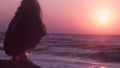 Beautiful girl watching pink sunset on the sea of dark color sitting on a stone