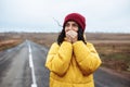 Beautiful girl wearing red hat and yellow jacket warms her hands with her breath on the lonely road. Young woman travels during