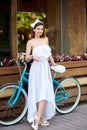 Beautiful girl in white dress posing with blue bike near a cafe. Royalty Free Stock Photo