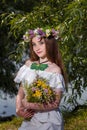 Girl in white dress and wreath holding field flowers bouquet on green willow background. 7th July, traditional slavic holiday with