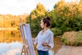 Beautiful girl, woman artist, summer by lake, river pond, draws picture, palette with paints, paintbrush white canvas