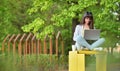 Beautiful girl works sitting with a laptop in a park on a background of green trees. Freelance work in a recreation area