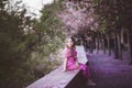 Beautiful girl 10 y.o. in pink dress sit in cherry blossom park on a spring day, flower petals falling from the tree