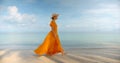 Beautiful girl in yellow long dress and hat walking at the edge of water and looking at the calm sea