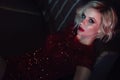 Beautiful glam blond model with provocative make up wearing red sequin dress relaxing on the sofa in night club