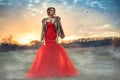 Beautiful glam model with updo hair wearing posh red fishtail dress and luxurious mink vest standing in the misty field at sunset Royalty Free Stock Photo