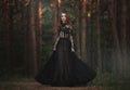 A beautiful gothic princess with pale skin and very long red hair in a black crown and a black long dress in a misty fairy forest.