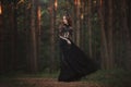 A beautiful gothic princess with pale skin and very long red hair in a black crown and a black long dress in a misty fairy forest.