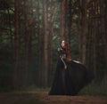 A beautiful gothic princess with pale skin and very long red hair in a black crown and a black long dress walks in a misty fairy-t Royalty Free Stock Photo