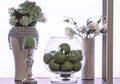 Beautiful shiny apples in a transparent glass vase and two vases with white flowers against a white glowing wall