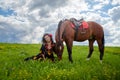 Beautiful gypsy girl sitting near horse in field with green glass in summer day and blue sky and white clouds background. Model in Royalty Free Stock Photo