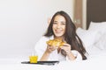 Beautiful happy Asian girl with long hair in white robe having breakfast Croissant and orange juice in the bed of hotel room Royalty Free Stock Photo